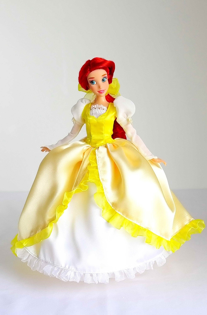 Pode incluir: Uma boneca de cabelo ruivo vestindo um vestido de cetim amarelo com acabamento de renda branca e um corpete branco. A boneca tem olhos azuis e um la&ccedil;o vermelho no cabelo.