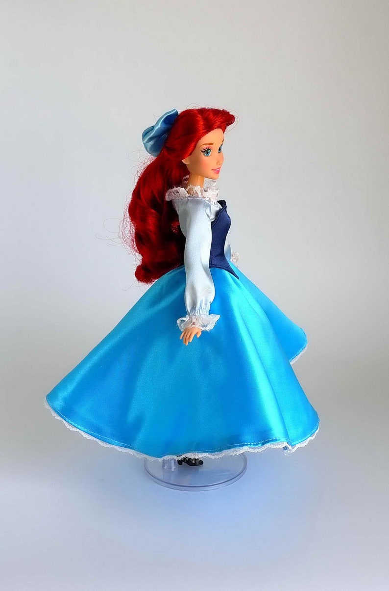 May include: A red-haired doll wearing a blue and white dress with a lace trim. The doll has a blue bow in her hair and is standing on a clear plastic stand.