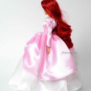 Peut inclure: Une poup&eacute;e aux longs cheveux roux, v&ecirc;tue d'une robe en satin rose avec une jupe blanche en dessous. La robe a des manches bouffantes et une jupe &agrave; plusieurs &eacute;paisseurs. La poup&eacute;e est pr&eacute;sent&eacute;e de profil sur fond blanc. Le texte "Malanedoll" est visible.