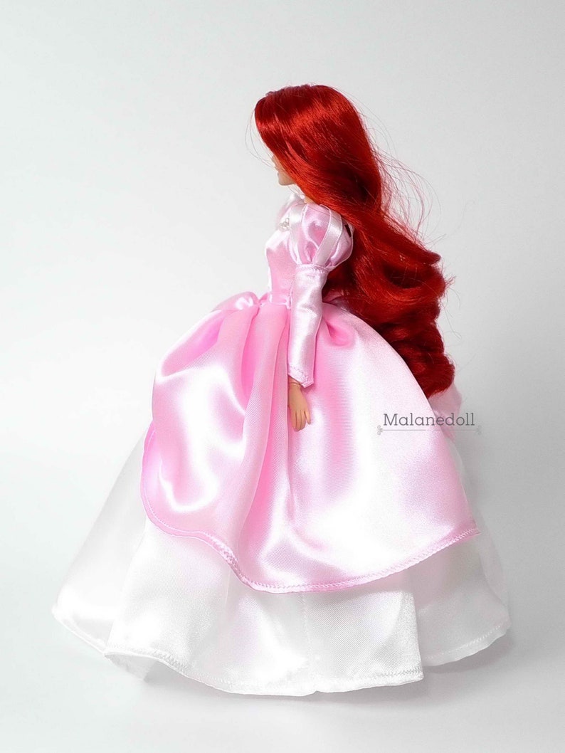 Peut inclure: Une poup&eacute;e aux longs cheveux roux, v&ecirc;tue d'une robe en satin rose avec une jupe blanche en dessous. La robe a des manches bouffantes et une jupe &agrave; plusieurs &eacute;paisseurs. La poup&eacute;e est pr&eacute;sent&eacute;e de profil sur fond blanc. Le texte "Malanedoll" est visible.