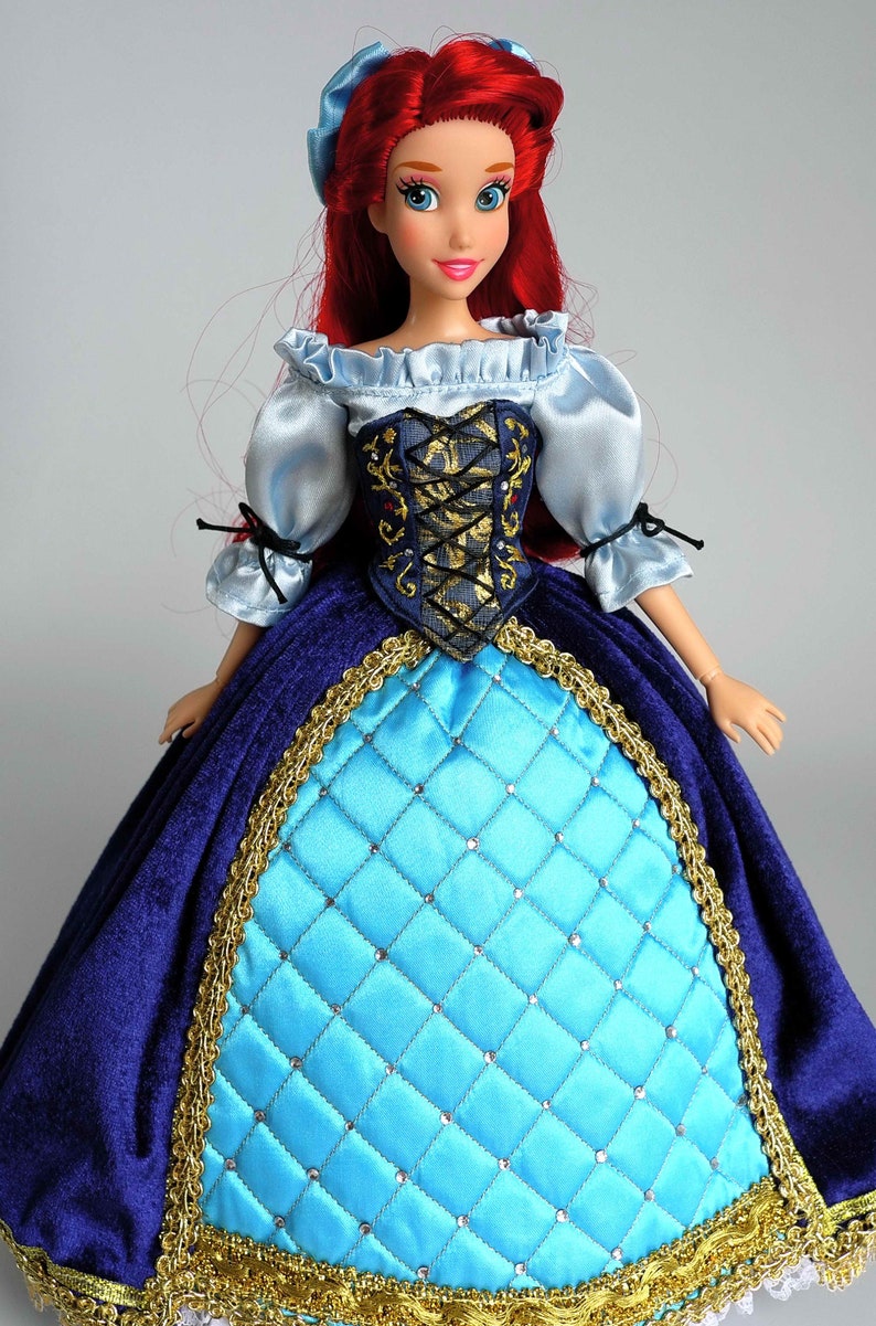 May include: A red-haired doll wearing a blue and gold dress with a blue bodice and a light blue, ruffled blouse. The doll has blue eyes and red lips.