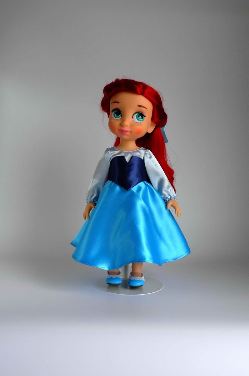 May include: A red-haired doll wearing a blue and white dress with a navy blue bodice. The doll has green eyes and is standing on a white stand.