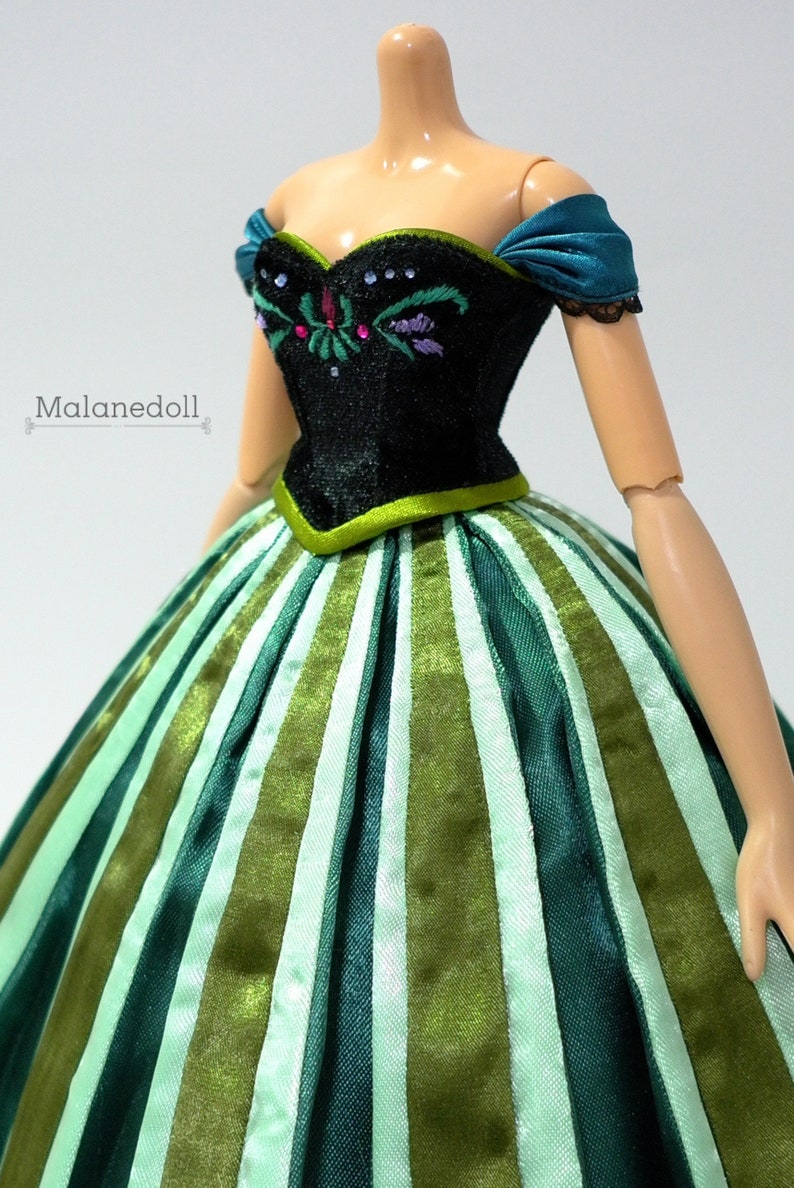 May include: A doll wearing a green and white striped dress with a black bodice featuring embroidered flowers. The doll has a teal sash and a black lace trim on the shoulders.