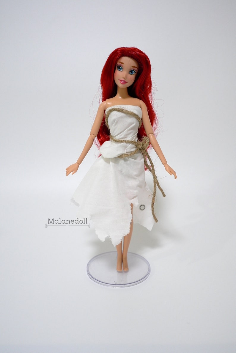 May include: A red-haired doll wearing a white dress with a brown rope belt. The doll is standing on a clear plastic stand.