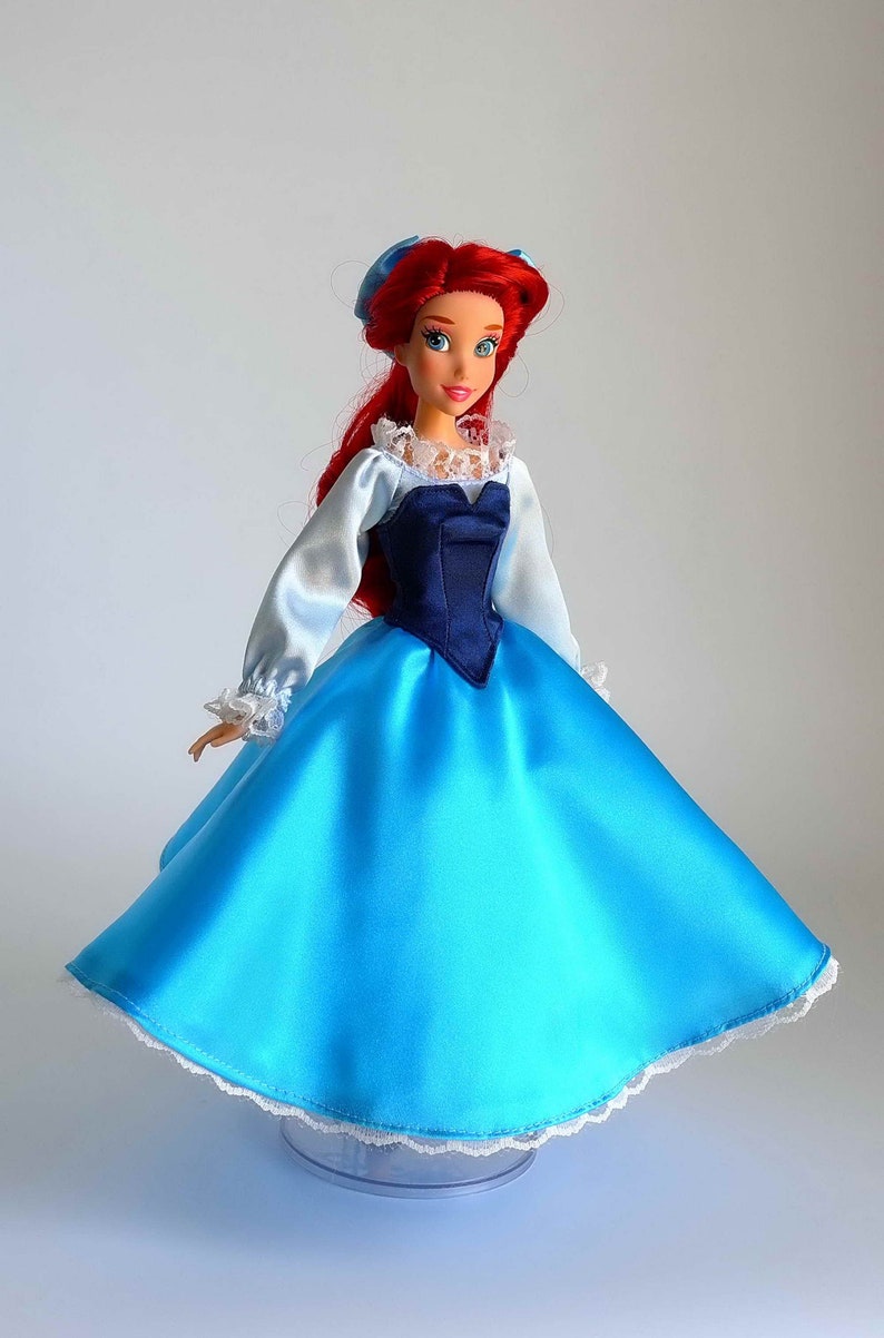 May include: A red-haired doll wearing a blue satin dress with a white lace trim and a navy blue bodice. The doll has blue eyes and is wearing a blue bow in her hair.