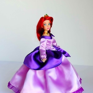 May include: A red-haired doll wearing a purple and pink satin ball gown with a gold crown. The doll has blue eyes and a green jewel necklace.
