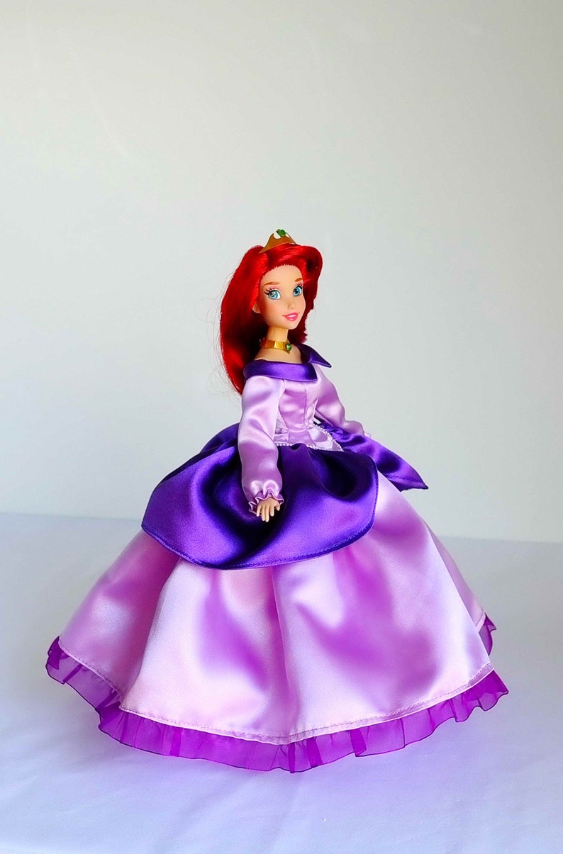 May include: A red-haired doll wearing a purple and pink satin ball gown with a gold crown. The doll has blue eyes and a green jewel necklace.