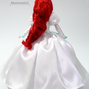 May include: A red-haired doll wearing a white satin wedding dress with a green trim. The doll is facing away from the camera.