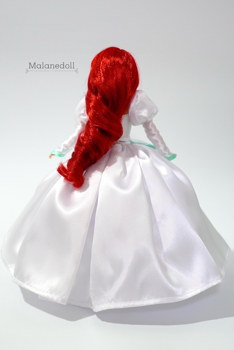 May include: A red-haired doll wearing a white satin wedding dress with a green trim. The doll is facing away from the camera.