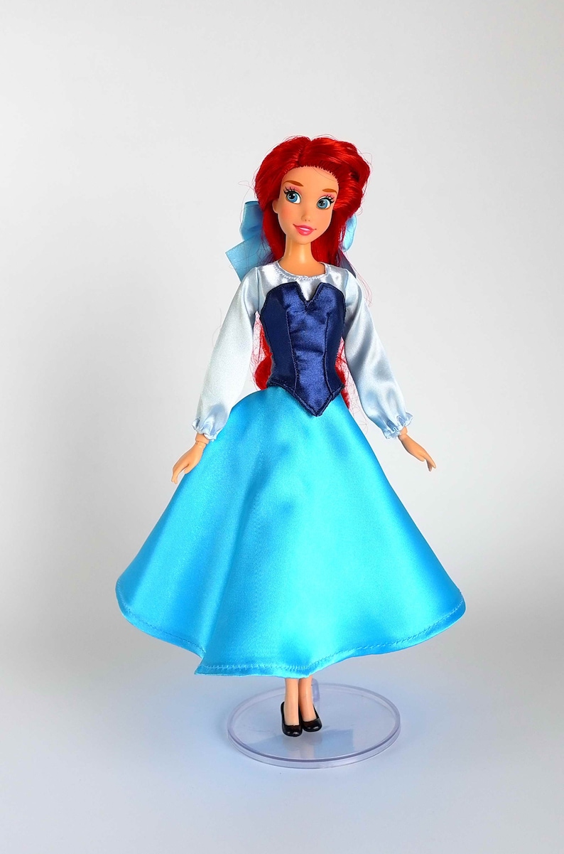 K&ouml;nnte beinhalten: Puppe einer Figur mit roten Haaren, die ein blaues Satinkleid und ein dunkelblaues Oberteil tr&auml;gt. Die Puppe hat eine hellblaue Schleife und schwarze Schuhe. Die Puppe steht auf einem durchsichtigen Kunststoffst&auml;nder.