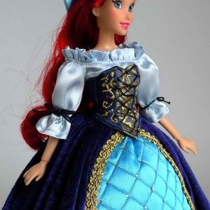 May include: A red-haired doll wearing a blue and gold dress with a blue bodice and a blue and white ruffled blouse. The doll has a blue headband and blue eyes.