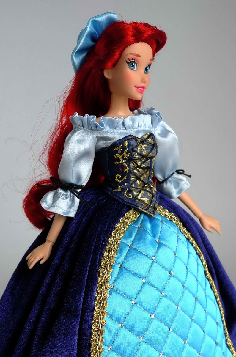 May include: A red-haired doll wearing a blue and gold dress with a blue bodice and a blue and white ruffled blouse. The doll has a blue headband and blue eyes.