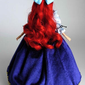 May include: A red-haired doll wearing a blue velvet dress with gold trim and a light blue bow in her hair.