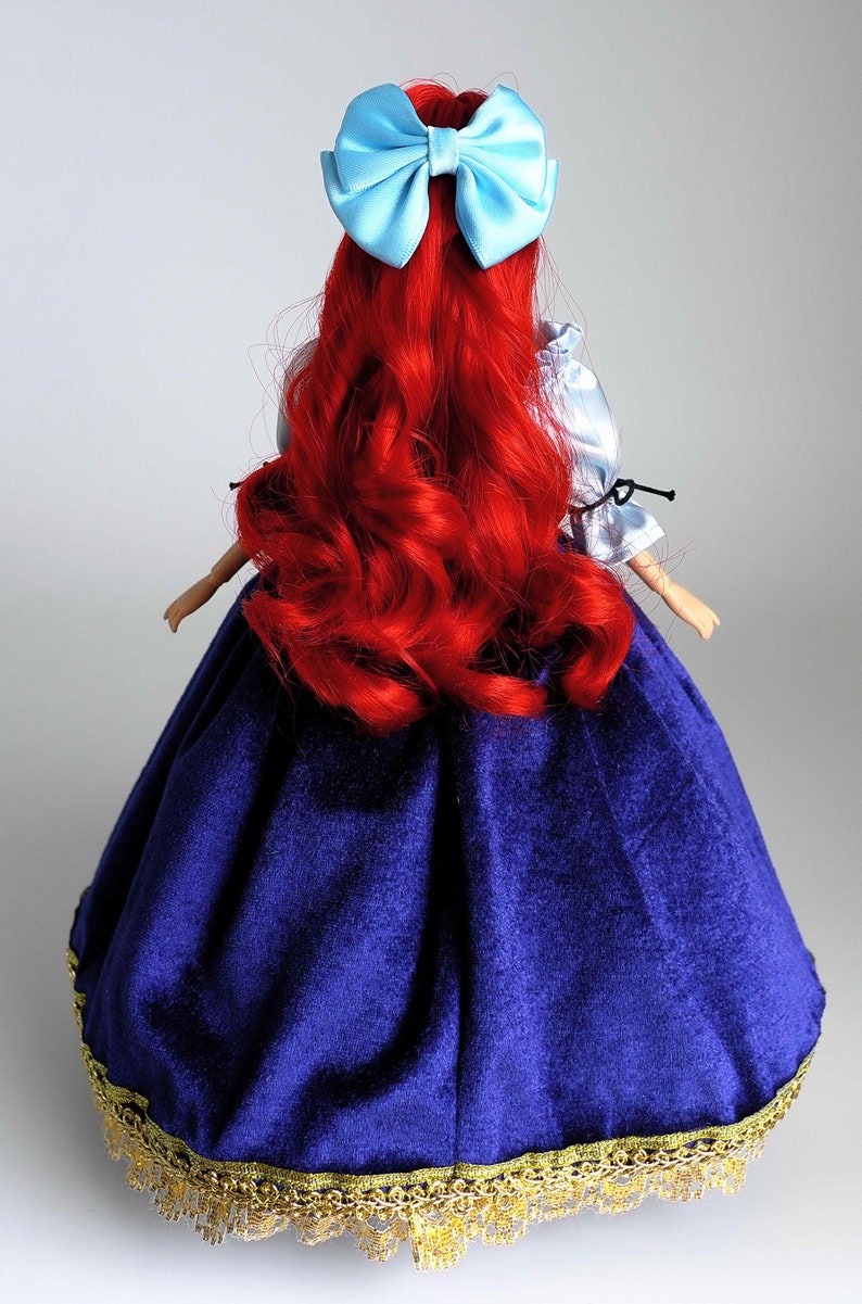 May include: A red-haired doll wearing a blue velvet dress with gold trim and a light blue bow in her hair.