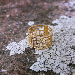 May include: A gold-colored ring with a geometric design. The ring is made of a thin metal and has a wide band.
