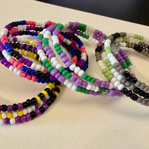 May include: Three spiral wrap bracelets made of small round glass beads. One bracelet features yellow, white, purple, and black beads. Another has blue, pink, purple, white, and green beads. The third bracelet consists of white, gray, black, and green beads.