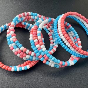 May include: A multi-strand coil bracelet made of small, round beads in alternating colors of light pink, white, and light blue. The beads are arranged in a repeating pattern, creating a flexible, spiraling design that wraps around the wrist.