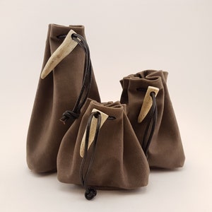 May include: Three brown suede drawstring pouches with antler toggles. The pouches are different sizes and are arranged in a row from largest to smallest.