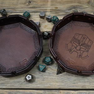 May include: Two brown leather dice trays with geometric designs and a helmet illustration. The trays are octagonal and surrounded by various multi-sided dice in green, silver, and black. The trays are on a wooden surface.