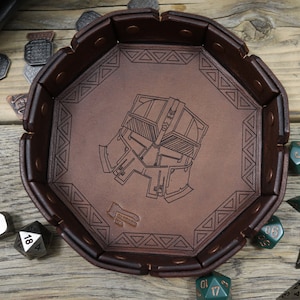 May include: A brown leather dice tray with a detailed engraved design of a helmet. The tray is octagonal and surrounded by a darker brown border. Various polyhedral dice in silver and green are scattered around the tray.