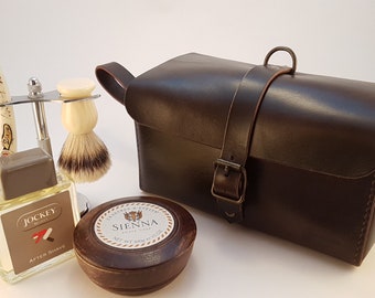 Pode incluir: Um kit de viagem de couro marrom com fecho de fivela, um pincel de barbear, uma navalha de barbear, uma garrafa de aftershave com o rótulo "Jockey After Shave" e um recipiente redondo de sabonete de barbear com o rótulo "Sienna Shaving Soap".