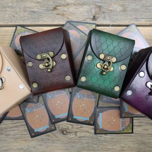 May include: Four rectangular leather deck boxes with metal latches, arranged in a row. The colors are natural tan, dark brown with a scale pattern, green with a scale pattern, and purple with a geometric line design, all resting on Magic cards.