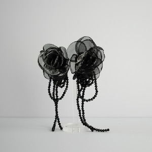 Op de afbeelding: Twee zwarte, roosvormige haaraccessoires met kralenaccenten. De rozen zijn gemaakt van gelaagde, doorschijnende stof met een glinsterend midden. Kralenstrengen hangen naar beneden en voegen elegantie toe. De accessoires worden op een transparante standaard gepresenteerd.