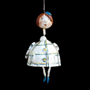 Può includere: Ornamento a forma di bambola in ceramica fatta a mano con cappotto bianco a quadri blu e gialli, testa marrone, cappello blu e scarpe blu. L'ornamento è appeso a un filo bianco su sfondo nero.