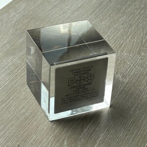 Puede incluir: Un bloque cuadrado de acrílico transparente con una placa metálica en el interior. La placa presenta el texto "Con licencia oficial de Cristóbal Colón" y "Jubileo del Quinto Centenario 1492-1992". El diseño es de Benno Welling.