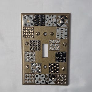 May include: A decorative light switch cover with a gold-colored metal finish. The cover features a grid of small, square tiles in black, white, and gold geometric patterns. The switch opening is centered.