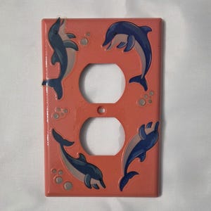 May include: Coral-colored electrical outlet cover featuring four blue and gray dolphins. The dolphins are arranged around the two outlet holes. Bubbles are also painted on the cover.