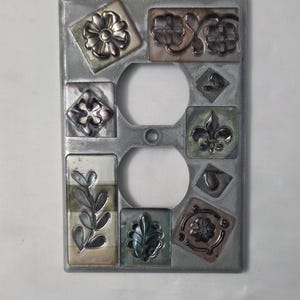 Può includere: Copertura decorativa argentata per presa elettrica con disegni floreali e di foglie in rilievo. La copertura presenta due aperture circolari per le prese ed è adornata da piastrelle quadrate e rettangolari con motivi botanici in rilievo.