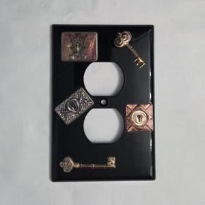 Peut inclure: Plaque de prise électrique noire avec des ornements métalliques décoratifs. Comprend une clé, une serrure et des motifs rectangulaires ornés dans des tons or, argent et cuivre. La plaque a deux ouvertures circulaires pour les prises.