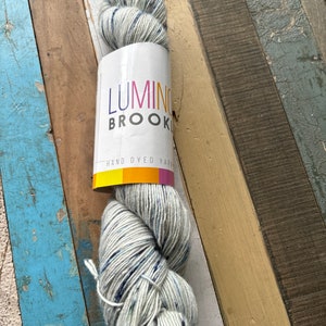 May include: A skein of hand-dyed yarn with a white label that reads "Luminous Brooklyn". The yarn is a light gray color with dark blue and white flecks. The yarn is sitting on a wooden surface with various colors.