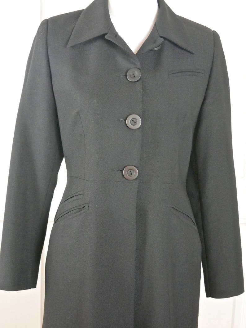 May include: A long black coat with a collar and five buttons down the front. The coat has pockets on the sides and a fitted waist.