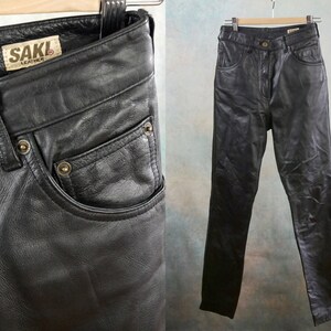 Könnte beinhalten: Schwarze Lederhose mit hoher Taille und geradem Bein. Die Hose hat einen Reißverschluss und einen Knopfverschluss. Das Etikett auf der Hose lautet "SAKI Leather".