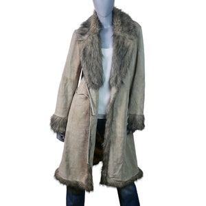 Op de afbeelding: Een lange, beige su&egrave;de jas met een kraag en bies van nep bont. De jas heeft een strikceintuur in de taille en een lange, vloeiende silhouet.
