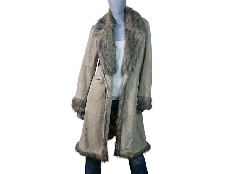 Op de afbeelding: Een lange, beige su&egrave;de jas met een kraag en bies van nep bont. De jas heeft een strikceintuur in de taille en een lange, vloeiende silhouet.