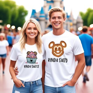 May include: A couple wearing white t-shirts with a Mickey Mouse design. The woman's shirt says "Kinda Sweet" and features a cupcake with Mickey Mouse ears. The man's shirt says "Kinda Salty" and features a pretzel Mickey Mouse.