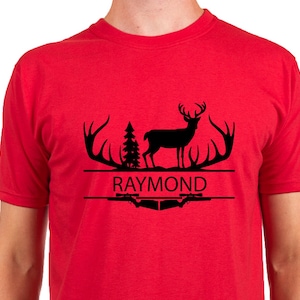 以下が含まれることがあります： 赤いTシャツに黒いグラフィックデザイン。デザインには、角のある鹿のシルエット、松の木、そして白い文字で「RAYMOND」という名前が含まれています。このシャツは半袖です。