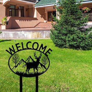 May include: Black metal welcome sign with a deer silhouette and mountain scene inside a circle. The sign says "WELCOME" in large letters.