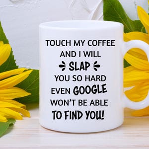 May include: White ceramic coffee mug with a curved handle. The mug features black text that reads: "TOUCH MY COFFEE AND I WILL SLAP YOU SO HARD EVEN GOOGLE WON'T BE ABLE TO FIND YOU!" Yellow sunflowers are in the background.
