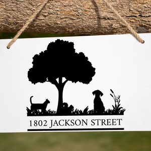 Puede incluir: Cartel rectangular blanco con una silueta negra de un árbol, gato, perro y hierba. El texto "1802 JACKSON STREET" está impreso en negro. El cartel está suspendido por una cuerda natural.