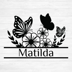 Op de afbeelding: Zwart-wit bloemen- en vlinderontwerp met de naam "Matilda" in een schreeflettertype. Het ontwerp toont drie vlinders, bloemen en bladeren, allemaal in silhouetstijl, tegen een witte houtnerf achtergrond.
