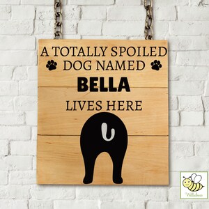 Könnte beinhalten: Holzschild mit dem Text "A TOTALLY SPOILED DOG NAMED BELLA LIVES HERE" in Schwarz. Das Schild zeigt eine schwarze Hunde-Schwanz-Grafik. Das Schild hängt an einer Kette vor einer weißen Backsteinmauer.