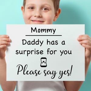 May include: A child holding a white sign with black text that reads "Mommy, Daddy has a surprise for you, Please say yes!" The sign includes a ring icon. The background is a solid light blue.