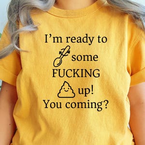 Peut inclure: T-shirt jaune moutarde avec des manches retroussées. Le texte noir dit "I'm ready to some fucking up! You coming?" avec une cuillère et un emoji caca. Le t-shirt est fait d'une matière douce.