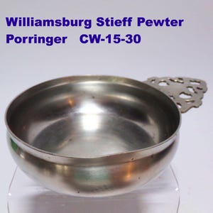May include: A silver pewter porringer with a decorative handle. The porringer is labeled "Williamsburg Stieff Pewter Porringer CW-15-30".