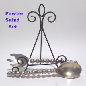 May include: Pewter salad set with a fork and spoon, displayed on a black metal stand. The fork and spoon have beaded handles. The text "Pewter Salad Set" is in purple.