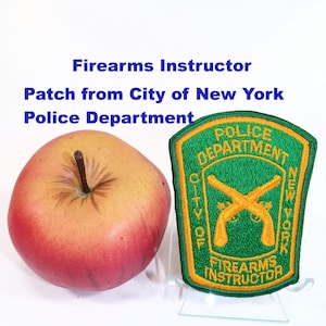 May include: A red and yellow apple next to a green and gold embroidered patch featuring the text Police Department City of New York Firearms Instructor with an image of two crossed flintlock pistols in the center of the shield-shaped patch.